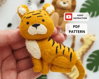 May include: A felt tiger toy with black stripes, white accents, and a cute face. The toy is being held in a hand. The image also includes text that says "VIDEO INSTRUCTION" and "PDF PATTERN".