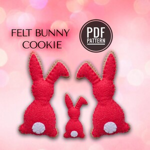 Peut inclure: Patron PDF pour biscuits de lapin en feutre. Trois lapins en feutre rouge avec des queues blanches, deux grands et un petit, sont représentés sur un fond rose.