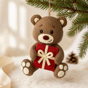 Pode incluir: Um enfeite de urso de peluche em feltro castanho pendurado por um fio. O urso tem um focinho cor de creme, nariz castanho escuro e olhos pretos. Segura um presente quadrado de feltro vermelho com uma fita e laço de feltro creme.