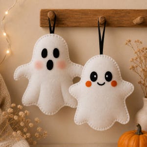 May include: Two white felt ghost ornaments hanging from a wooden rack. The left ghost has black oval eyes, an oval mouth, and pink cheeks. The right ghost has round black eyes, a small curved smile, and orange circular cheeks. Both have black ribbons.