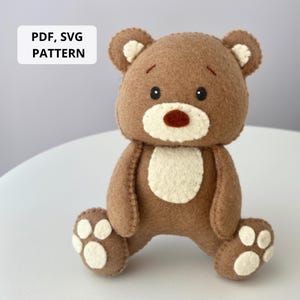 May include: A handmade brown felt teddy bear with a white belly and paw pads. The bear has a dark red nose and black eyes. The text "PDF, SVG PATTERN" is in a white rectangle in the upper left corner.