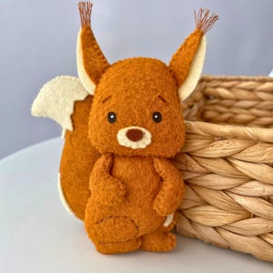 May include: A handmade felt squirrel figurine in shades of orange, cream, and brown. The squirrel has large ears, a fluffy tail, and a sweet expression. The figurine is next to a woven basket.