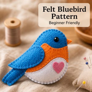 May include: A handmade felt bluebird in shades of blue, orange, and white. The bird features a pink heart and a black bead eye. The text "Felt Bluebird Pattern Beginner Friendly" is displayed in the upper right corner.