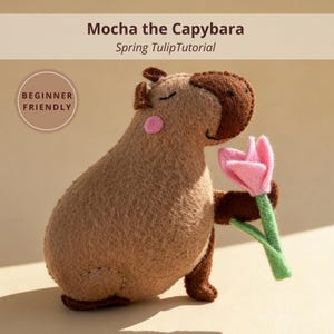 May include: A handmade felt capybara toy in shades of brown, holding a pink felt tulip. The toy has a round pink cheek and closed eyes. Text reads "Mocha the Capybara Spring Tulip Tutorial" and "Beginner Friendly".