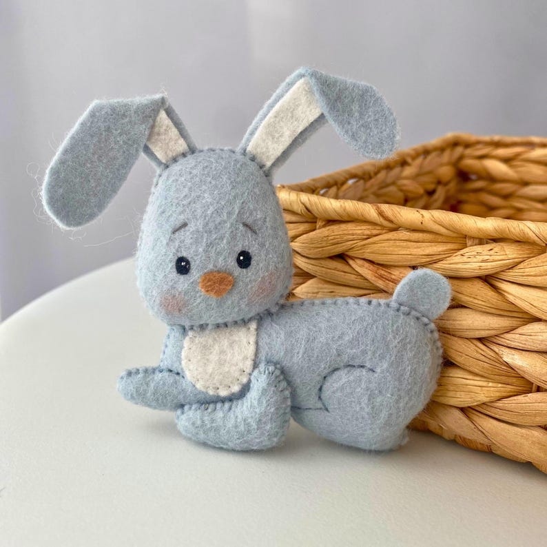 May include: A handmade light blue felt bunny toy with long ears, a white belly, and a small tail. The bunny has black eyes, a peach nose, and pink cheeks. It is positioned in front of a woven basket.