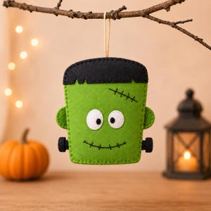 May include: A felt hanging ornament shaped like a green monster head with black hair, white eyes with black pupils, a stitched mouth, and black bolts on the sides. The ornament is suspended by a thin tan string from a brown branch.