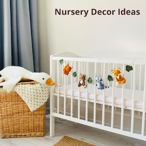 May include: A white crib with a garland of felt woodland animals, including a squirrel, bear, raccoon, and fox. A white goose plush toy rests on a wicker basket. The text "Nursery Decor Ideas" is at the top.