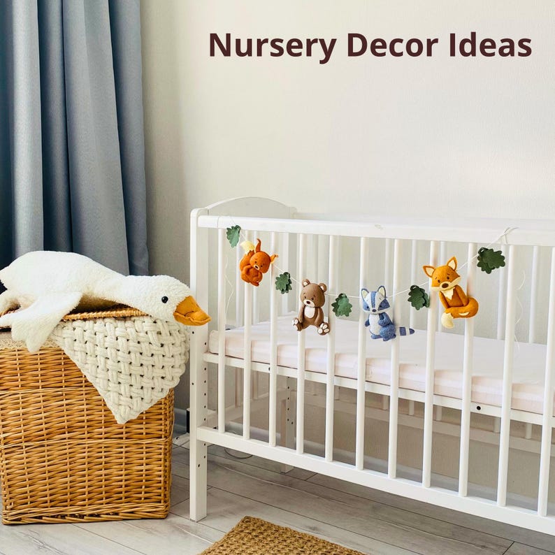 May include: A white crib with a garland of felt woodland animals, including a squirrel, bear, raccoon, and fox. A white goose plush toy rests on a wicker basket. The text "Nursery Decor Ideas" is at the top.