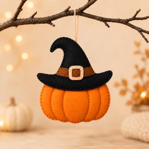 May include: A felt hanging ornament shaped like an orange pumpkin wearing a black witch hat. The hat features a brown band with a beige buckle. The pumpkin has visible stitched edges and is suspended by a thin tan string from a dark branch.