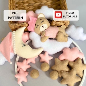 May include: A pink and brown felt baby mobile with a sleeping bear, a crescent moon, clouds, and stars. The mobile is hanging from a white wooden hoop. The text "PDF PATTERN" and "VIDEO TUTORIALS" are visible in the image.