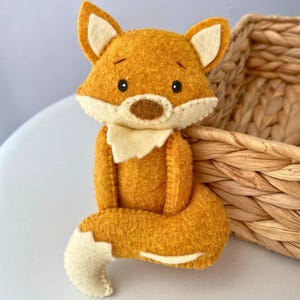 May include: A handmade felt fox figurine in shades of orange, cream, and brown. The fox has a white chest, ears, and tail tip, with black eyes and a brown nose. The fox is sitting, with its tail curled around its body. A woven basket is in the background.