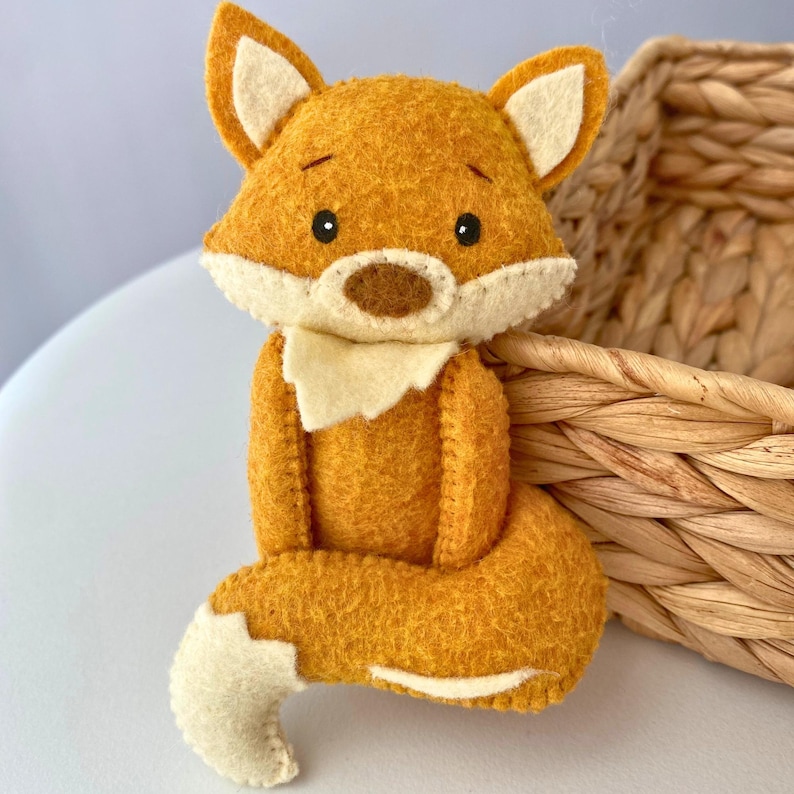 May include: A handmade felt fox figurine in shades of orange, cream, and brown. The fox has a white chest, ears, and tail tip, with black eyes and a brown nose. The fox is sitting, with its tail curled around its body. A woven basket is in the background.