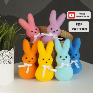 Peut inclure: Cinq lapins en peluche de Pâques en feutre de différentes couleurs : rose, orange, jaune, bleu et bleu clair. Chaque lapin a un ruban blanc noué autour du cou et des yeux en boutons noirs. Les lapins sont disposés en demi-cercle sur une surface blanche. Le texte « VIDEO INSTRUCTION » et « PDF PATTERN » est visible sur l’image.
