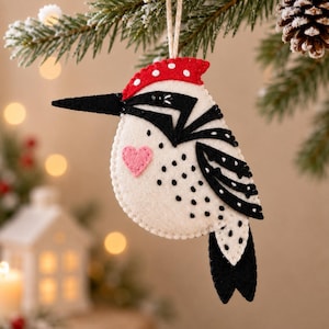 May include: A felt bird ornament featuring a cream-coloured body with black wing details, a long black beak, and a red cap with white polka dots. A small pink heart is stitched onto the chest, and the ornament hangs from a simple twine loop.