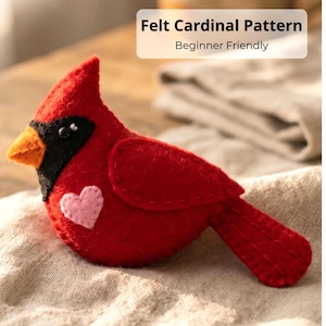 May include: A handcrafted red felt cardinal bird with a black mask, orange beak, and a pink heart on its chest. The text "Felt Cardinal Pattern Beginner Friendly" is displayed above the bird.