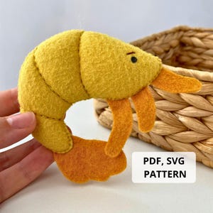 May include: A handmade felt shrimp toy in shades of yellow and orange. The shrimp has a curved body, orange legs, and small black eyes. The text "PDF, SVG PATTERN" is in a white rectangle.