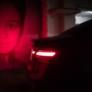 May include: A black car with red taillights parked in a dimly lit garage. The car is partially obscured by a red poster of a person's face.