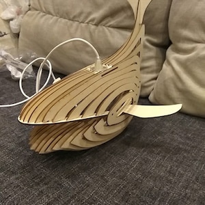 Könnte beinhalten: Ein Lampenschirm aus Holz in Form eines Wals mit einem weißen Kabel, das von ihm herabhängt. Der Lampenschirm steht auf einem grauen Sofa.