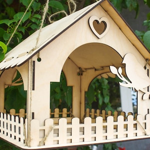 Puede incluir: Un comedero para pájaros de madera con forma de casa con un corazón recortado en el techo. El comedero tiene una pequeña valla alrededor de la parte inferior y una silueta de pájaro en el lateral. Está colgado de una cuerda.