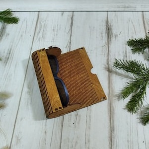 May include: A wooden box with a hinged lid, designed to hold sunglasses. The box is made of light brown wood with a striped pattern. The sunglasses are black and are partially visible inside the box.