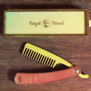 Può includere: Un pettine da barba in legno e una scatola di legno con la scritta "Royal Beard" sono esposti su una superficie di legno. Il pettine è giallo e rosso, simile a un rasoio diritto. La scatola è di colore chiaro con una cornice in legno scuro.