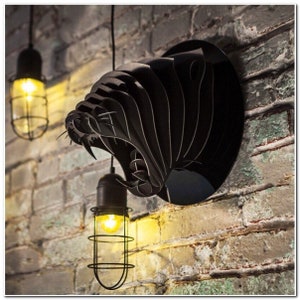 Puede incluir: Una escultura de pared de metal negro de la cabeza de un animal con dientes afilados, montada en una pared de ladrillo. Dos bombillas enjauladas cuelgan del techo sobre la escultura.