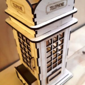 May include: A wooden model of a red telephone booth with the word "TELEPHONE" printed on the top. The model has a small drawer on the bottom and a window with a grid pattern.