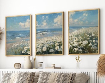 May include: Set of three framed landscape paintings. Each features a beach scene with a blue sky, white clouds, and white flowers. The frames are a light wood color, adding a natural touch to the artwork.