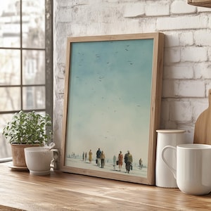 May include: A framed art print featuring a beach scene with silhouetted figures walking along the shore under a light blue sky with birds. The artwork is in a light wooden frame, displayed on a wooden surface.