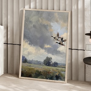 Könnte beinhalten: Ein gerahmtes Landschaftsgemälde zeigt eine Gruppe von Gänsen im Flug vor einem bewölkten Himmel. Das Kunstwerk zeigt ein Feld in Grün- und Brauntönen mit Bäumen in der Ferne. Das Gemälde ist in einem hellfarbigen Holzrahmen eingefasst.