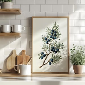 Puede incluir: Una impresión botánica enmarcada que presenta una ilustración detallada de una rama de olivo con hojas verdes y aceitunas azul oscuro. La obra de arte está sobre un fondo crema, enmarcada en madera clara. La impresión se exhibe en una encimera de cocina.