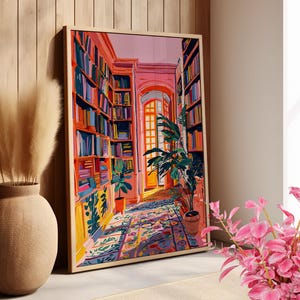 Op de afbeelding: Een ingelijste kunstdruk toont een kleurrijk bibliotheekinterieur. Boekenkasten vol boeken in verschillende kleuren staan langs de muren. Een patroonkleed leidt naar een deuropening, met potplanten. Het kunstwerk zit in een houten lijst.