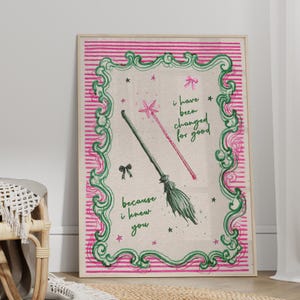 Pode incluir: Uma impressão emoldurada com um design caprichoso. A obra de arte apresenta uma varinha rosa e uma vassoura verde, com o texto "i have been changed for good" e "because i knew you". O fundo tem listras rosa e uma borda decorativa verde.