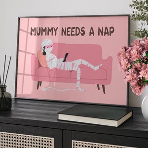 May include: Framed art print featuring a cartoon mummy relaxing on a pink sofa, holding a phone and remote control. The text "MUMMY NEEDS A NAP" is displayed above the mummy. The artwork is set against a pink background.
