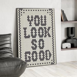 May include: Framed wall art with the words "YOU LOOK SO GOOD" in dark gray letters. The letters are made of small, hexagonal tiles, set against a background of off-white hexagonal tiles. The frame is black.
