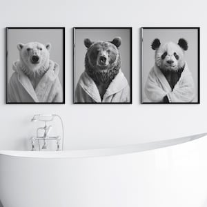 May include: Three black and white framed photographs of bears wearing white bathrobes. The bears are a polar bear, a brown bear, and a panda bear. The photographs are hanging on a white wall above a white bathtub.