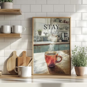 Könnte beinhalten: Ein gerahmter Druck mit den Worten "STAY awhile" über einem Aquarellbild einer Küchenszene. Eine rote Kaffeetasse steht auf einem Holztisch. Weiße Tassen und Schneidebretter befinden sich auf einem Regal.