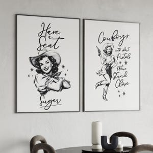 May include: Two framed black and white prints with vintage-style illustrations. One features a woman in a cowboy hat with the text "Have a Seat Sugar." The other shows a woman with a gun and the text "Cowboys with short pistols please stand close."