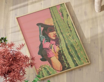 May include: Framed art print featuring a cowgirl with a revolver, partially obscured by a cactus. The artwork has a pink background and a vintage aesthetic. The cowgirl wears a hat, scarf, and gloves, holding the gun. The frame is a light wood color.