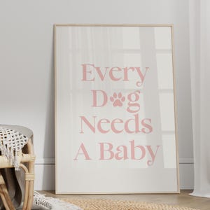 Op de afbeelding: Een ingelijste print met de tekst "Every Dog Needs A Baby" in roze letters. De print heeft een witte achtergrond en een licht houten frame. De afbeelding leunt tegen een muur in de buurt van een rieten stoel.