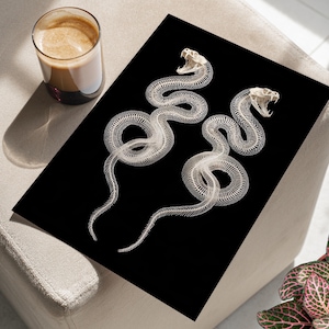 Może przedstawiać: Czarny nadruk przedstawiający dwa białe szkielety węży z otwartymi pyskami. Szkielety są szczegółowo dopracowane, z widocznymi żebrami i kręgami. Nadruk znajduje się na czarnym tle, tworząc uderzający kontrast. Filiżanka kawy znajduje się w lewym górnym rogu.