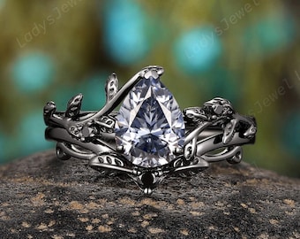 Könnte beinhalten: Ein schwarzer Verlobungsring mit einem birnenförmigen, klaren Edelstein. Der Ring hat ein florales Design mit Blättern und Ranken. Der Ring ist auf einem dunklen, strukturierten Stein platziert, mit einem verschwommenen grünen und blauen Hintergrund.