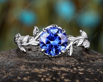 May include: A silver ring with a blue sapphire gemstone set in a delicate floral design. The ring features two branches with leaves on either side of the stone.