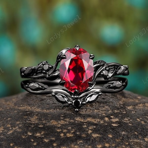 May include: A black gold engagement ring set with a large red oval gemstone and black diamond accents. The ring features a delicate vine design.