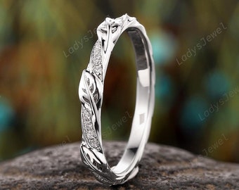 May include: A delicate silver wedding band with a vine design and small diamonds. The band is set against a blurred background of green foliage.