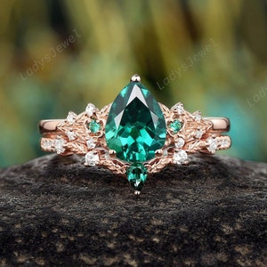 May include: A pear-shaped emerald engagement ring set in rose gold with a delicate floral design and smaller emerald accents. The ring is set on a gray stone background.