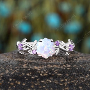May include: A silver ring featuring a hexagonal opal center stone. The ring is adorned with small purple gemstones and leaf-like details. The ring is set on a dark, textured surface.