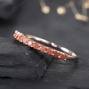 May include: A rose gold band ring with a row of small, round, orange-red gemstones. The ring is set on a dark gray, textured surface.