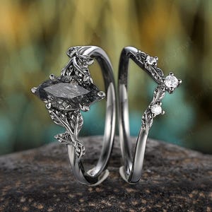 May include: A two-piece ring set featuring a dark metal band with a marquise-cut black rutilated quartz center stone and leaf-shaped accents, paired with a matching curved band adorned with three small, clear, round-cut accent stones.