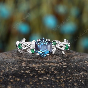 Könnte beinhalten: Ein silberner Ring mit einem blauen sechseckigen Edelstein, der von kleineren grünen Edelsteinen umgeben ist. Der Ring hat ein zartes, blattförmiges Design.